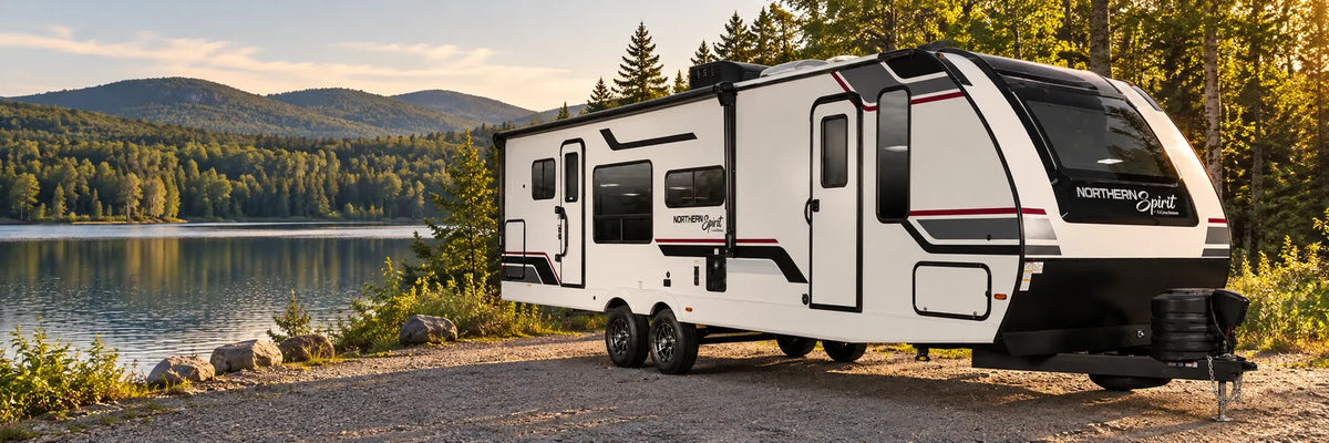Roulotte Northern Spirit de Coachmen au bord d’un lac, devant des collines boisées.